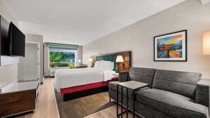 Una habitación de hotel moderna con cama, sofá, televisor y ventana con vistas a zonas verdes.