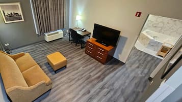 Habitación de hotel con sofá, escritorio con lámpara, televisión, baño pequeño con espejo y lavamanos, y una fotografía de la ciudad en la pared.