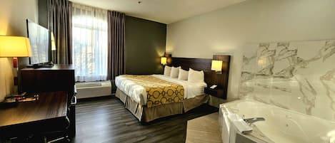 A hotel room with a bed, a desk, a TV, a desk lamp, a wall-mounted lamp, a large mirror, and a bathtub.