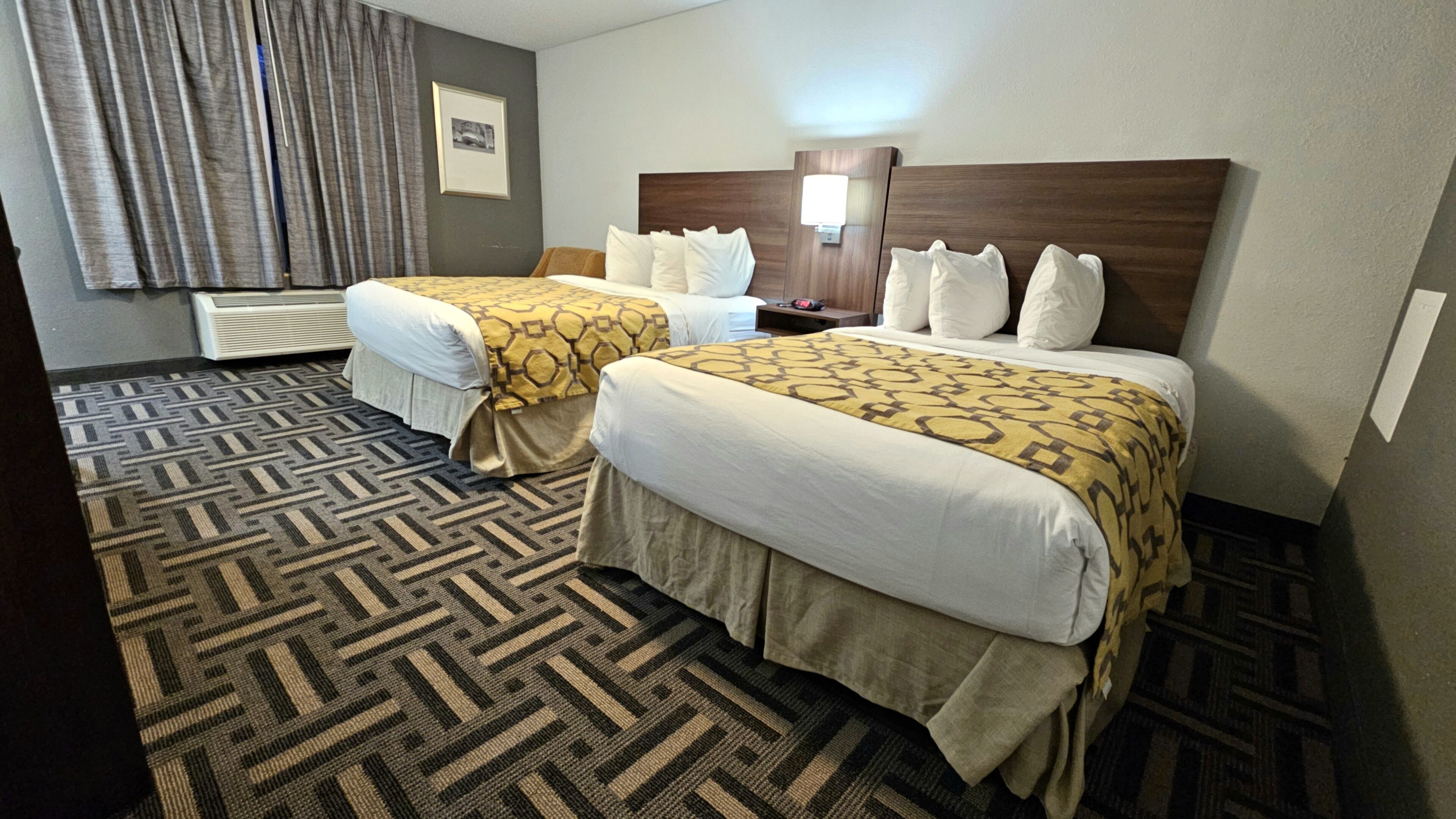 Quarto de hotel com duas camas, carpete estampado, quadro na parede e mesa de cabeceira com abajur.