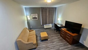 Habitación amoblada con sofá, reposapiés, televisión, escritorio, silla, lámpara, equipo de aire acondicionado y un cuadro en la pared.