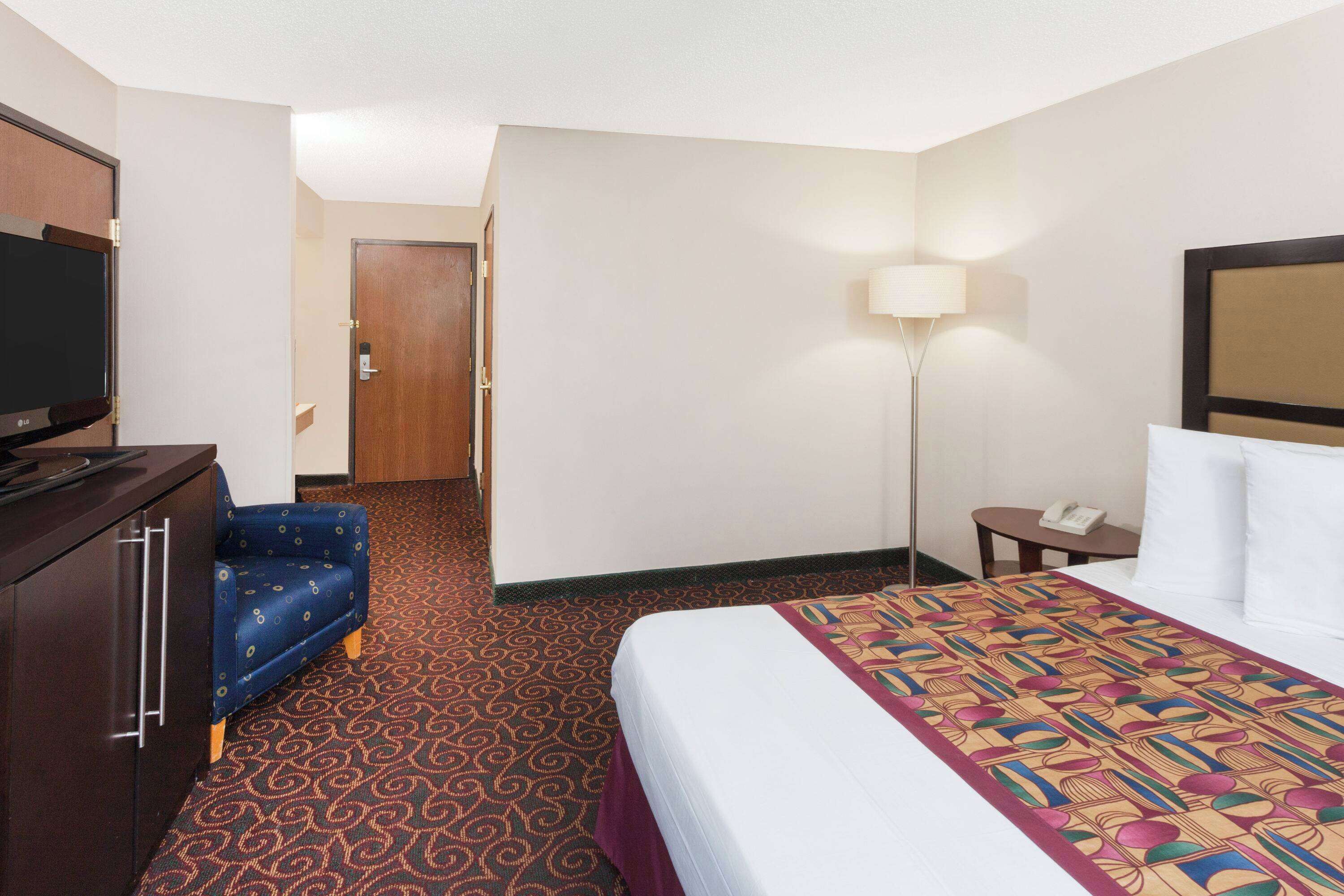 A hotel room with a bed, a television on a dresser, a desk with a lamp, and a door leading to another room.