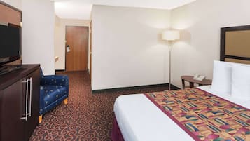 A hotel room with a bed, a television on a dresser, a desk with a lamp, and a door leading to another room.