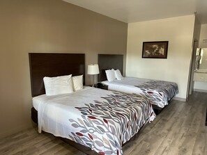 Habitación de hotel con dos camas, una mesita de noche con lámpara, un cuadro enmarcado en la pared y piso de madera.
