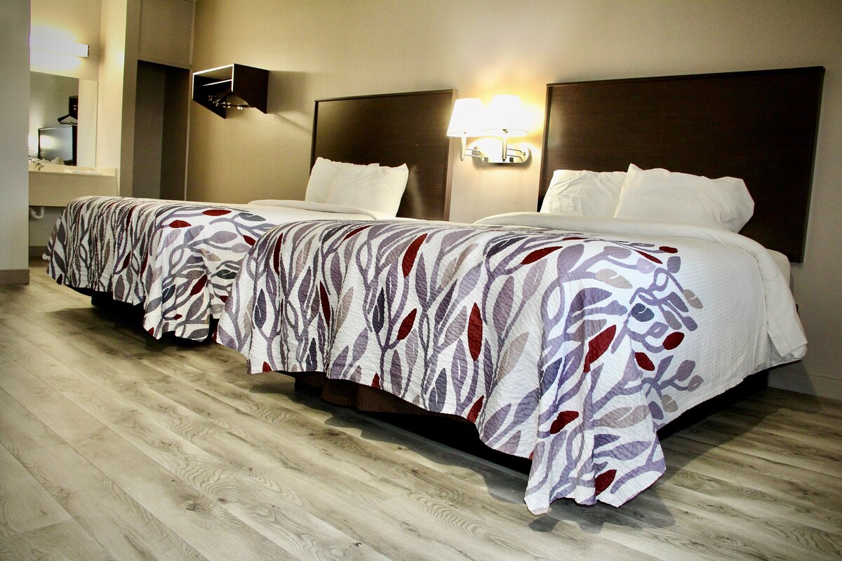 A hotel room with two beds, each with patterned bedspreads, a nightstand, a wall-mounted lamp, and a wooden floor.
