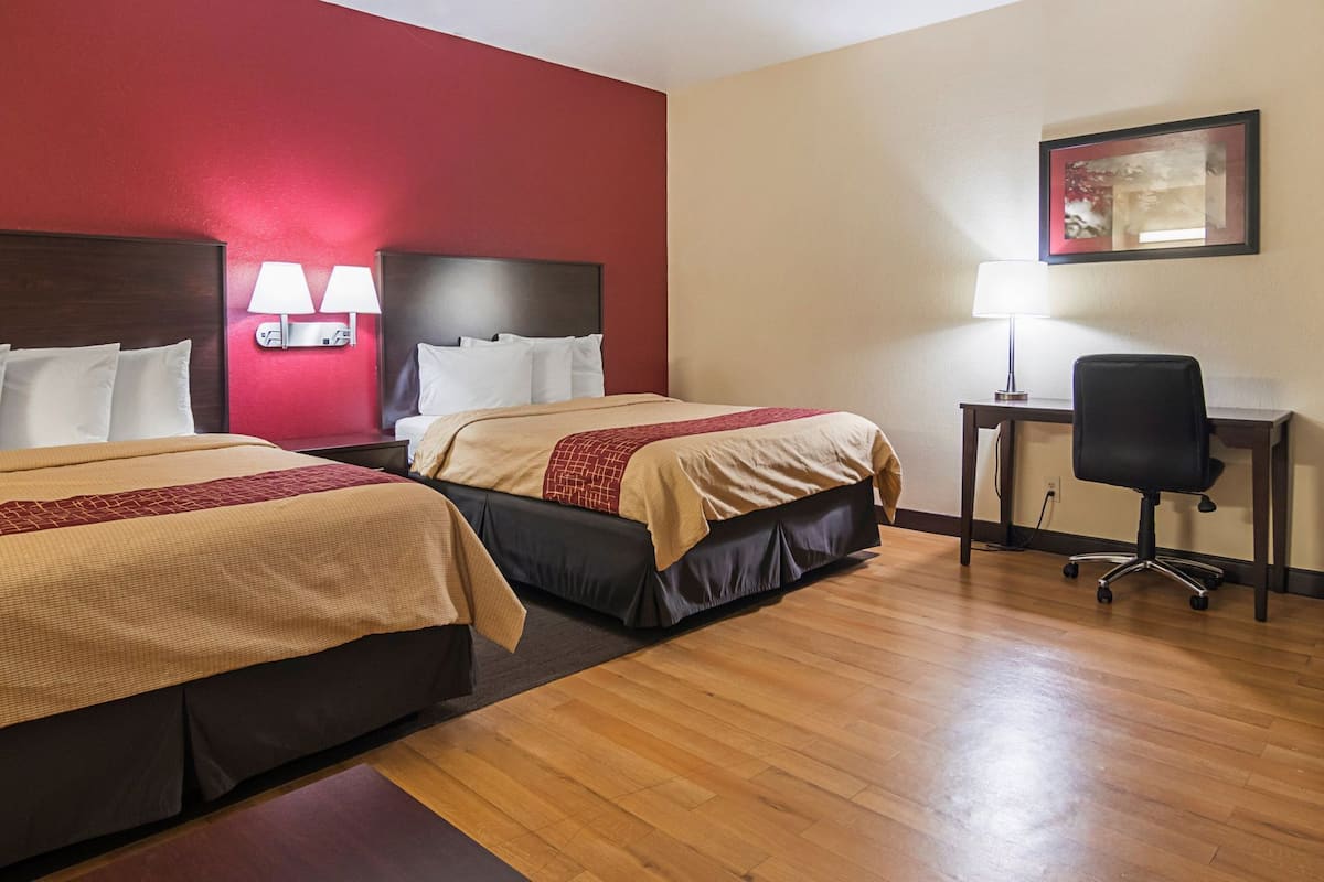 A hotel room with two beds, a desk, a chair, and a wall-mounted lamp.