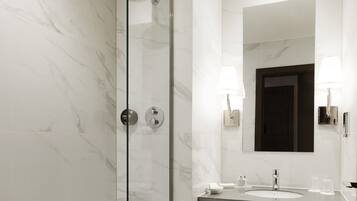 Un baño moderno con ducha envuelta en vidrio, pared con azulejos de mármol, un espejo grande con luces empotradas y un lavabo con encimera oscura.