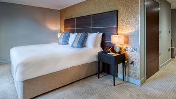 A hotel room with a large bed, a nightstand with a lamp, a telephone, and a wall-mounted control panel.