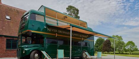 A green double-decker bus with "NEVER ORDINARY ALWAYS ALDWARK" written on the side, parked on a gravel surface with two wooden beach chairs in front.