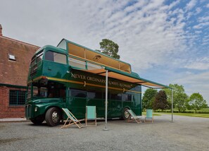 A green double-decker bus with "NEVER ORDINARY ALWAYS ALDWARK" written on the side, parked on a gravel surface with two wooden beach chairs in front.