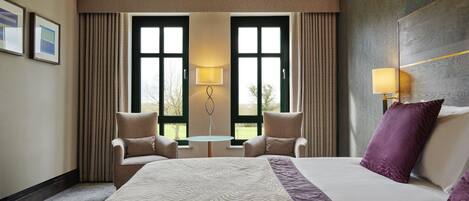 Habitación de hotel con una cama grande, dos sillones, una mesita y vistas amplias a través de ventanas grandes.