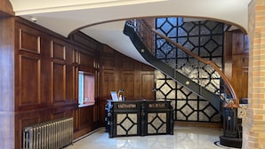 A grand foyer with a polished marble floor, a wooden reception desk, a radiator, and a staircase with a wrought iron railing. - Aldwark Manor Estate (York)
