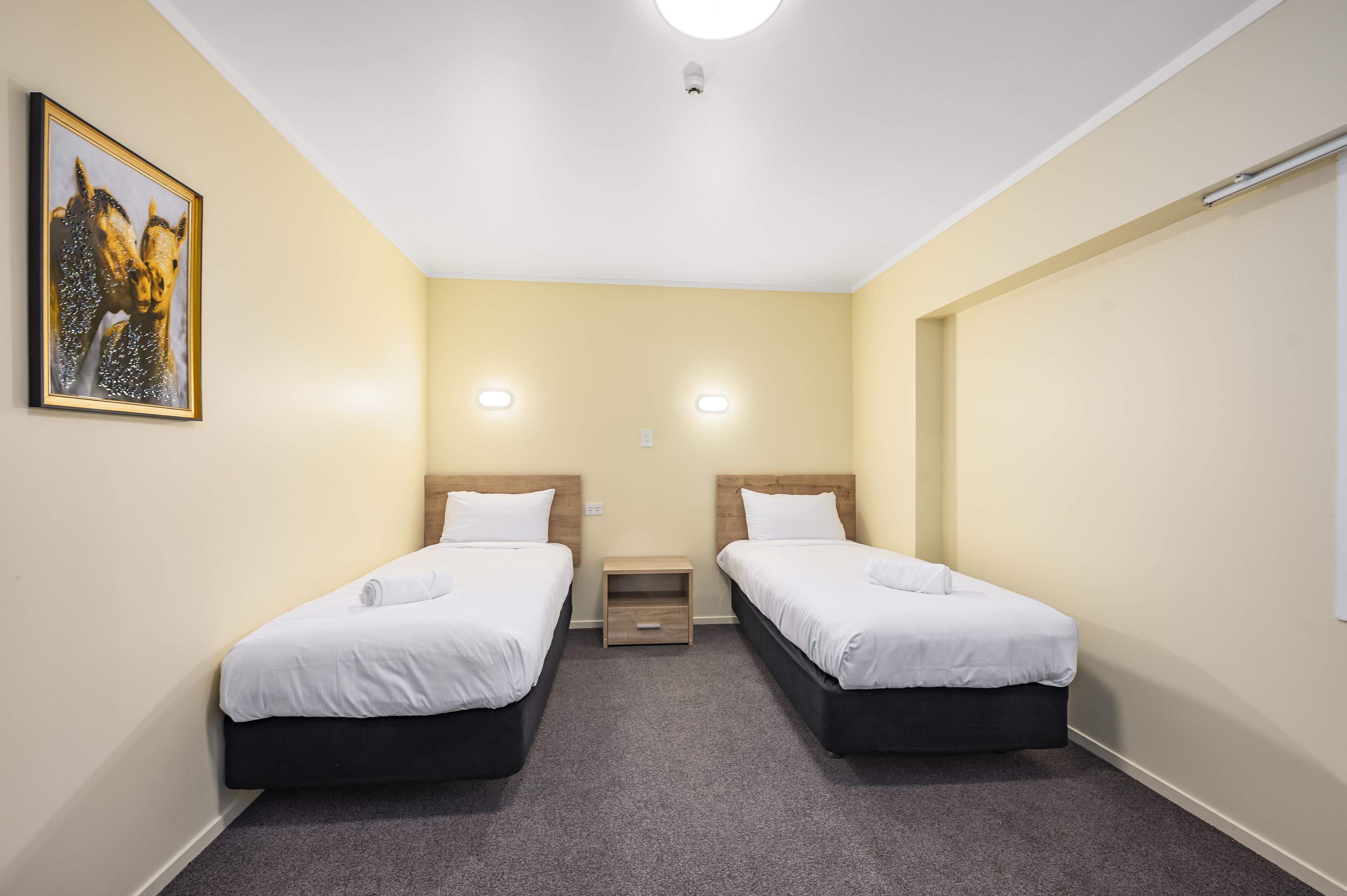 A small, clean room with two single beds, a small table, and a painting on the wall.