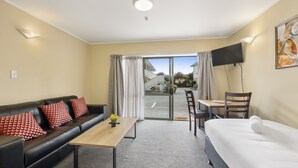 A hotel room with a black leather sofa, a wooden coffee table, a white bed, a dining table with chairs, and a TV mounted on the wall. - Fenton Court Motel (Rotorua)