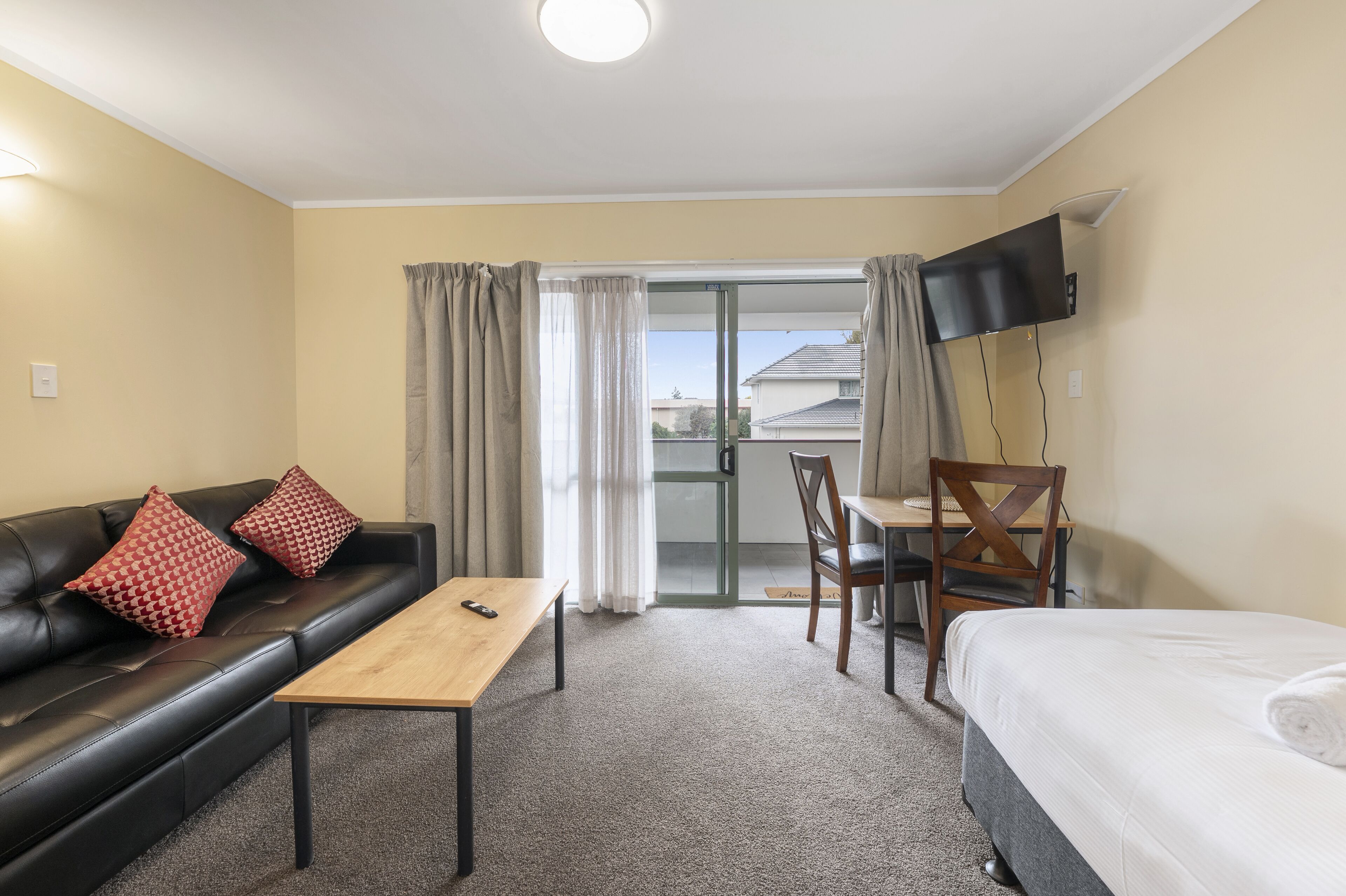 Ein Hotelzimmer mit schwarzem Ledersofa, einem Holztisch, einem Bett, einem Schreibtisch mit Stuhl, einem an der Wand befestigten Fernseher und einer Glastür, die auf einen Balkon führt.