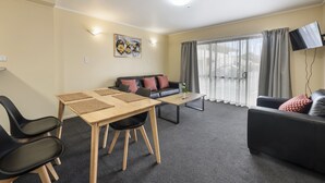 A modern living room with a sofa, a dining table with chairs, a flat-screen TV mounted on the wall, and a large window with curtains. - Fenton Court Motel (Rotorua)