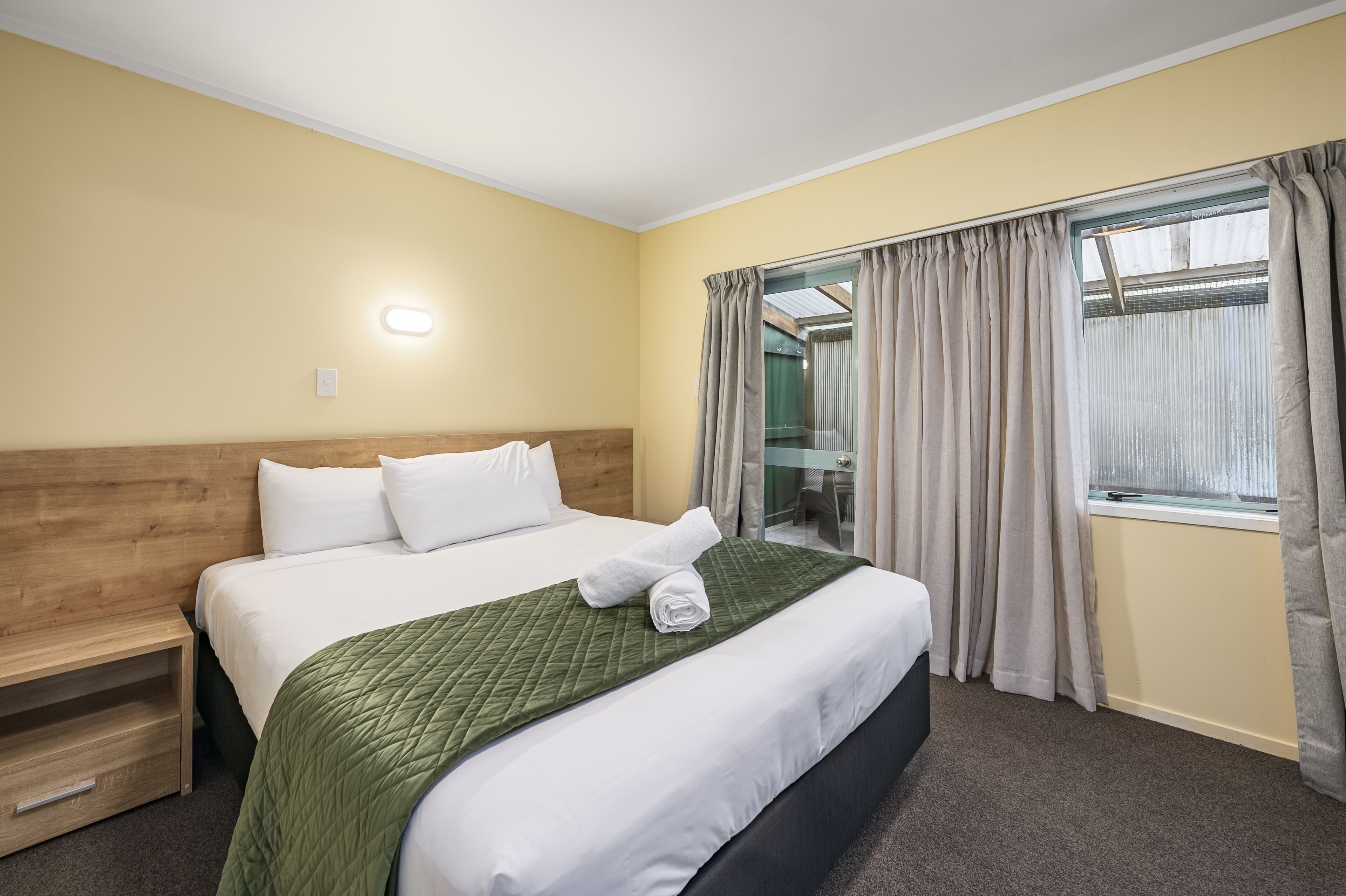 Habitación de hotel con cama, cabecera de madera, colcha verde acolchada y ventana con cortinas.