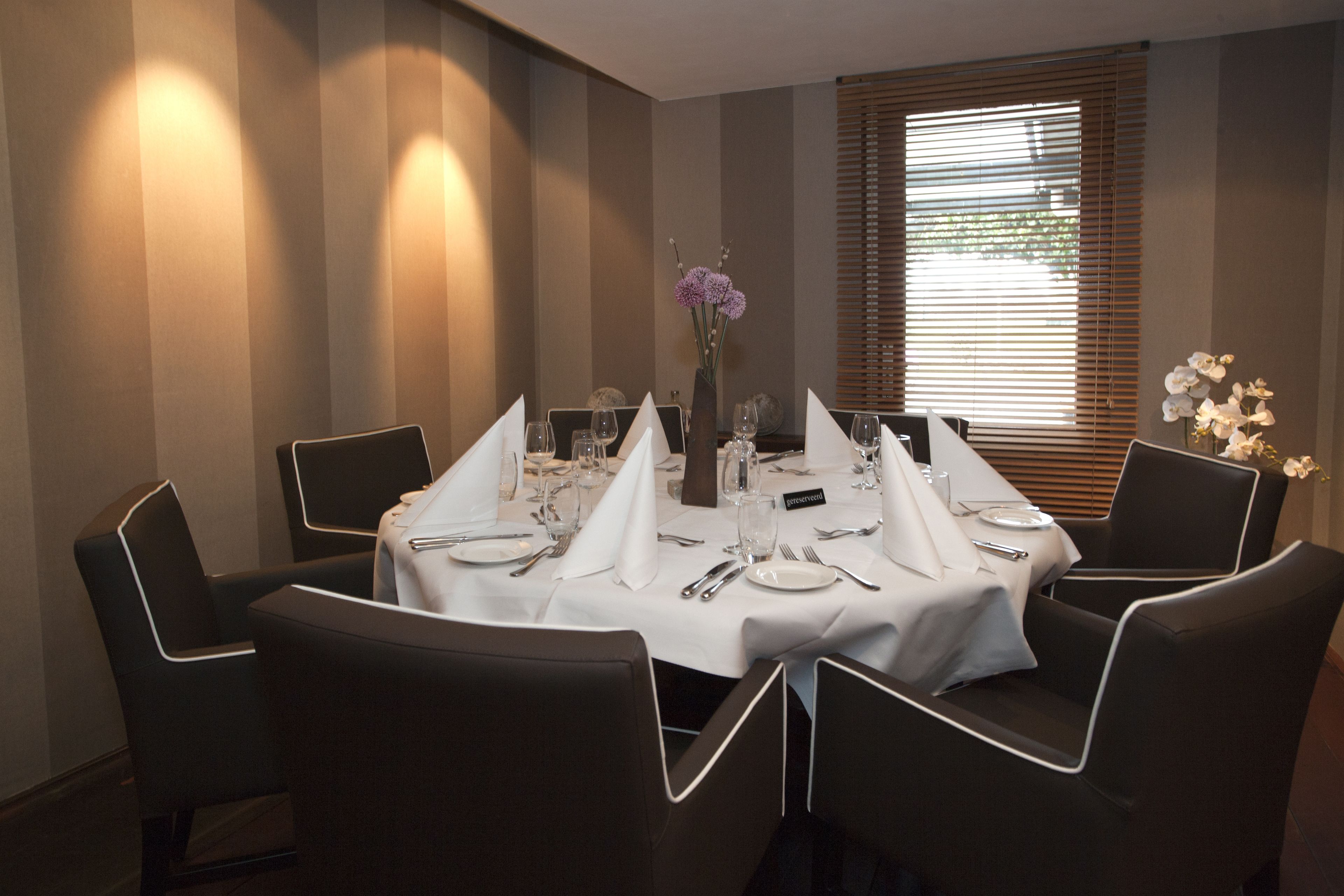 Un coin repas avec une table ronde dressée pour quatre personnes, entourée de chaises noires à l’assise blanche. Un vase rempli de fleurs est posé sur la table, et une fenêtre avec des stores se trouve à l’arrière-plan.