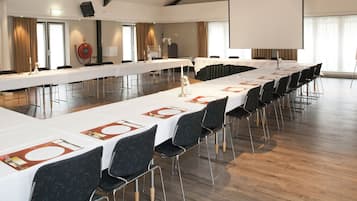 Une salle de conférence avec de longues tables dressées pour un repas, des chaises et un écran de projection.