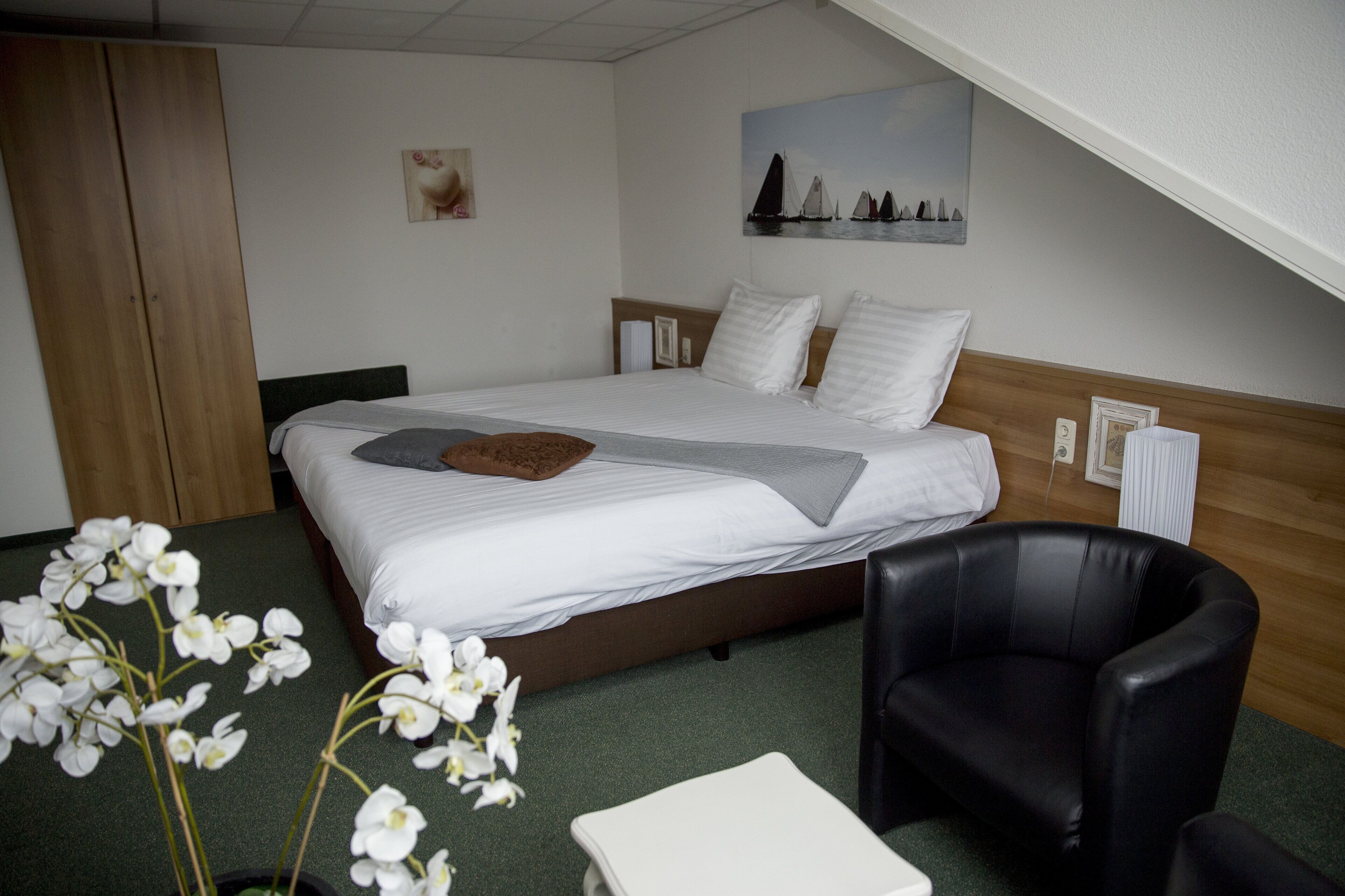 Une chambre d’hôtel avec un lit, un fauteuil en cuir noir, une petite table et des orchidées blanches posées sur le sol.