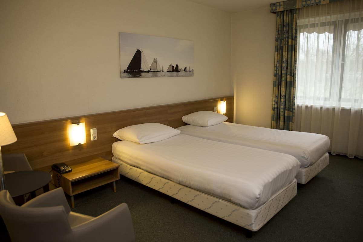 Habitación de hotel con dos camas, un escritorio con lámpara, una silla, un cuadro en la pared y una ventana con cortinas.