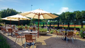 Un área de estar al aire libre con mesas y sillas, grandes sombrillas y un campo de béisbol al fondo.