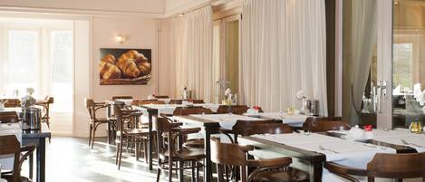 A dining area with multiple tables set for service, wooden chairs, and a large window with curtains.