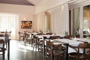 A dining area with multiple tables set for service, wooden chairs, and a large window with curtains.