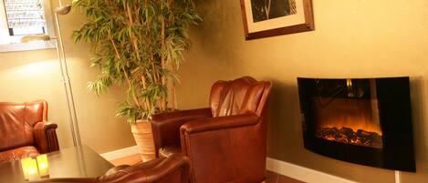 A cozy living room with leather chairs, a glass coffee table, a large potted plant, a wall-mounted flat-screen TV, and a fireplace.