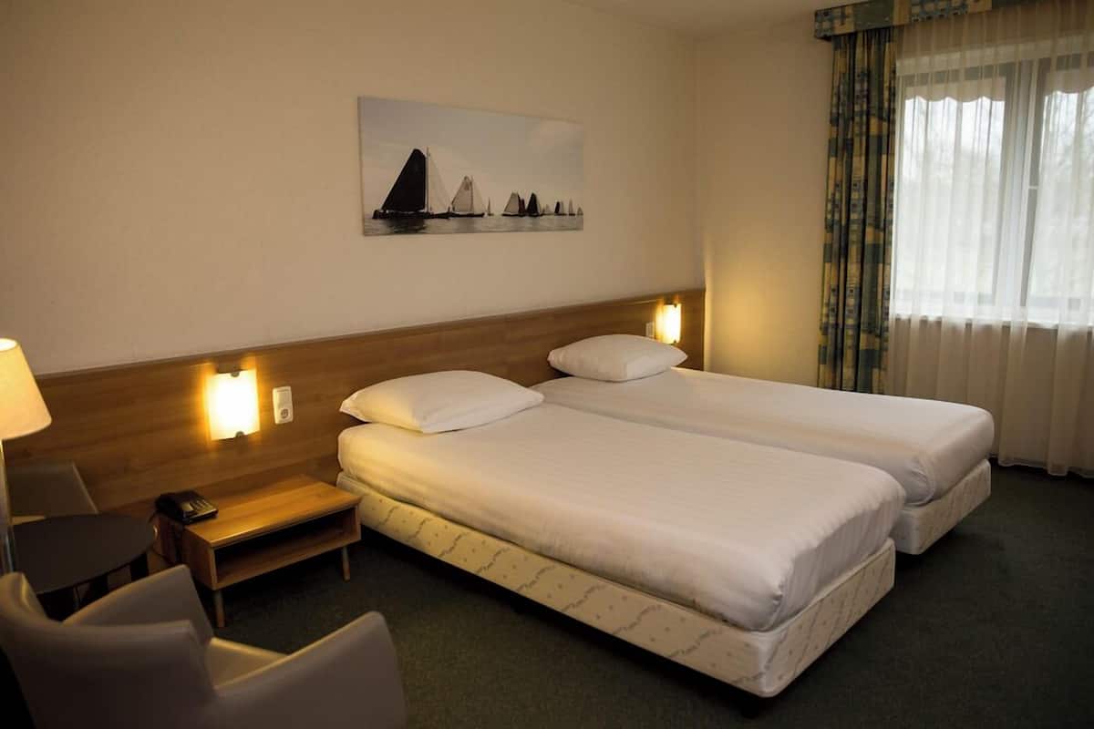 Habitación de hotel con dos camas, un escritorio, una silla, una mesita de noche y un cuadro en la pared.