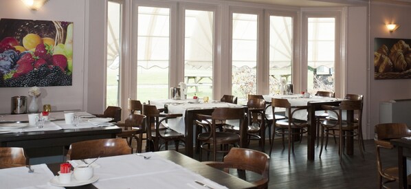 A dining area with multiple tables and chairs, large windows, and artwork on the walls.
