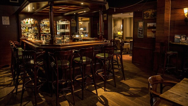 Una zona de bar con mostrador de madera, varios taburetes y una amplia variedad de botellas y vasos.