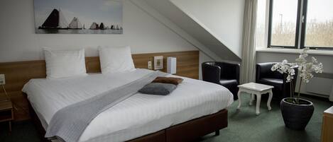 Habitación de hotel con una cama grande, una mesita pequeña, una silla, una planta en maceta y un cuadro en la pared.