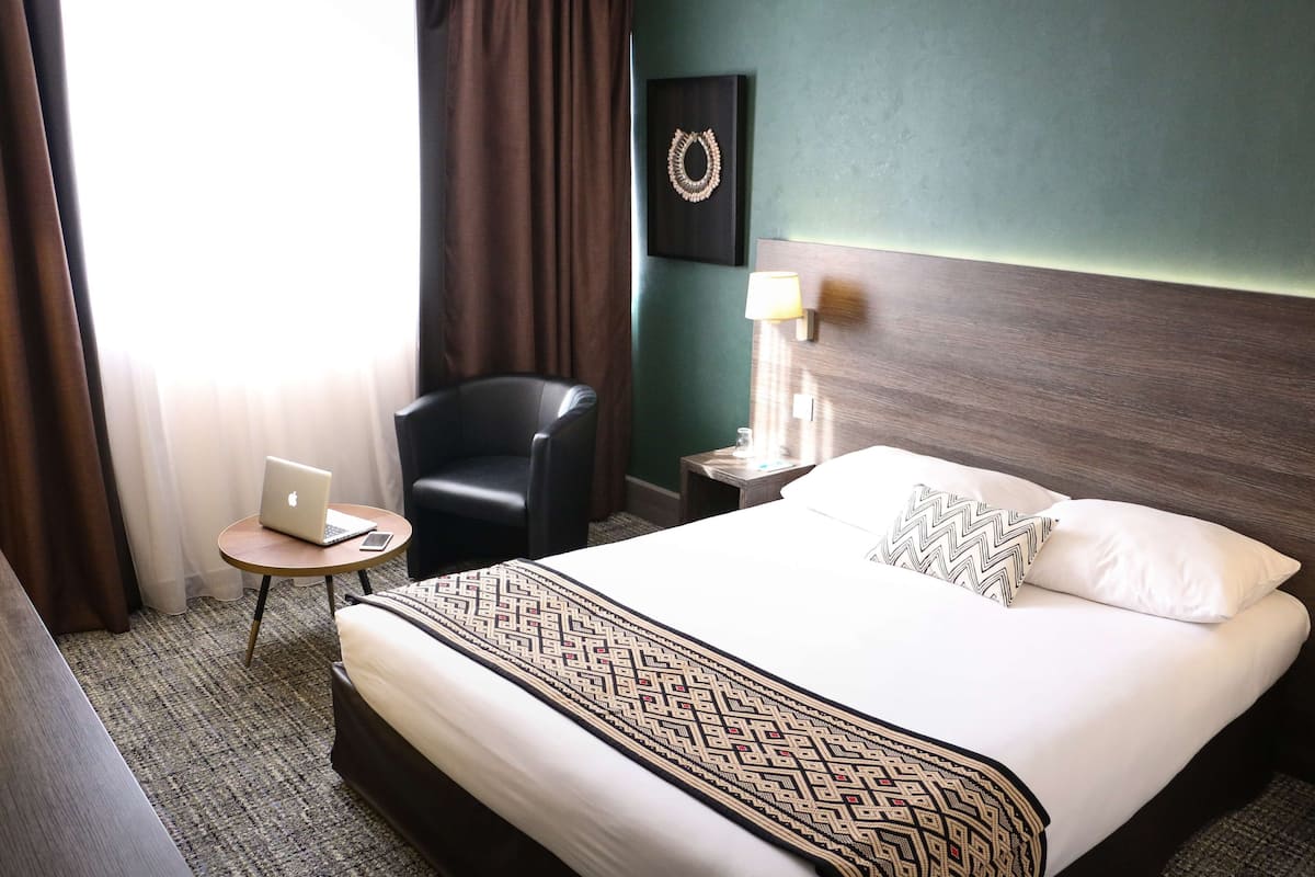 A hotel room with a bed, a chair, a small table with a laptop, a lamp, and a wall decoration.