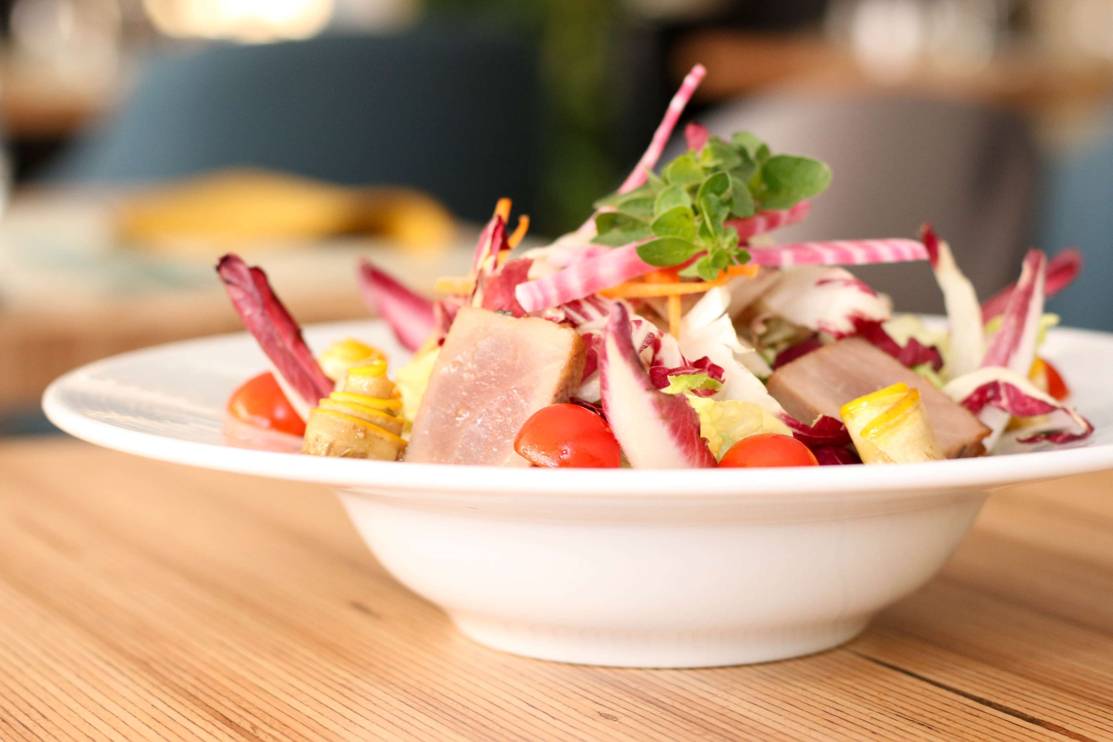 A white bowl containing a salad with various vegetables and meat, placed on a wooden table.