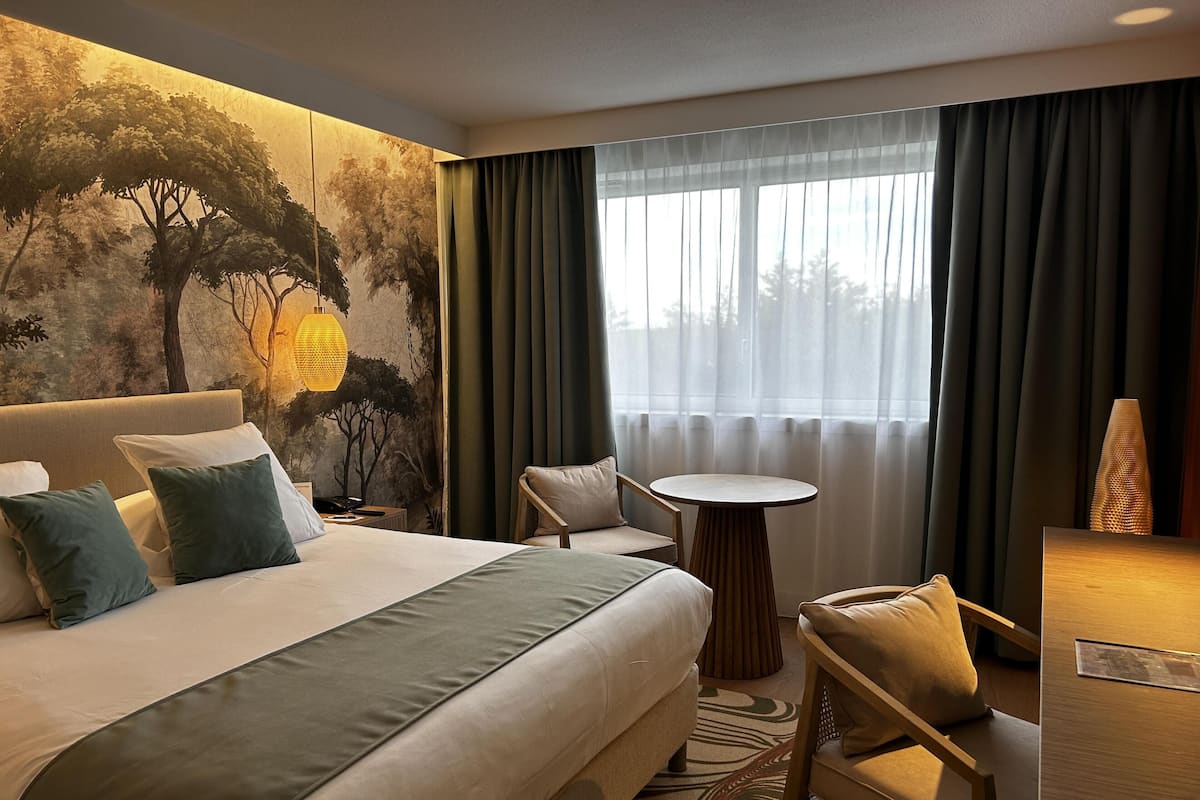 A hotel room with a large bed, a small round table, a chair, a desk, and a mural on the wall.