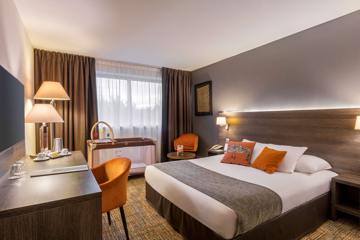 A hotel room with a bed, desk, chair, and a small table with a lamp.