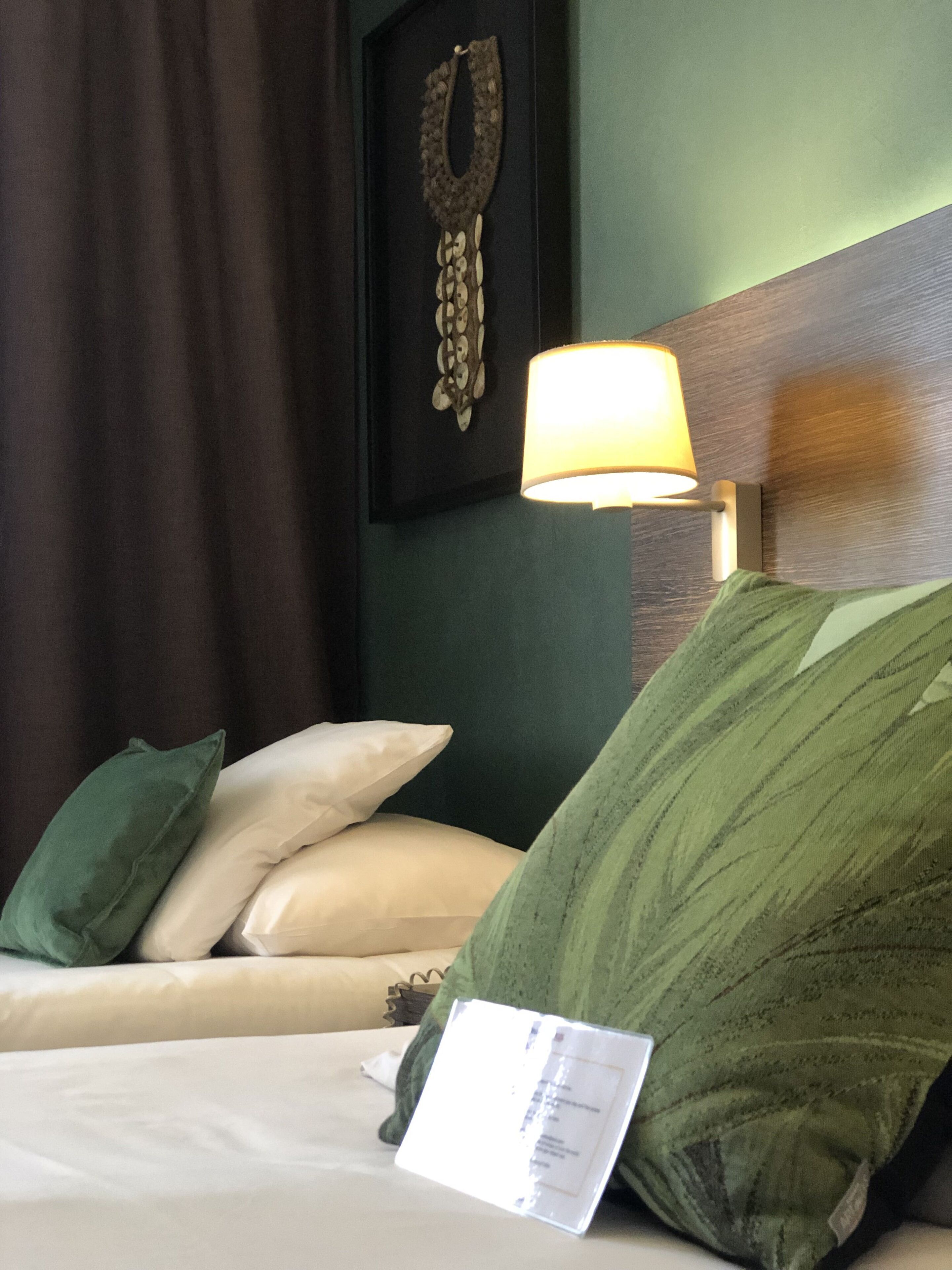 A hotel room with a bed, green and white pillows, a wall-mounted lamp, and a decorative piece above the bed.
