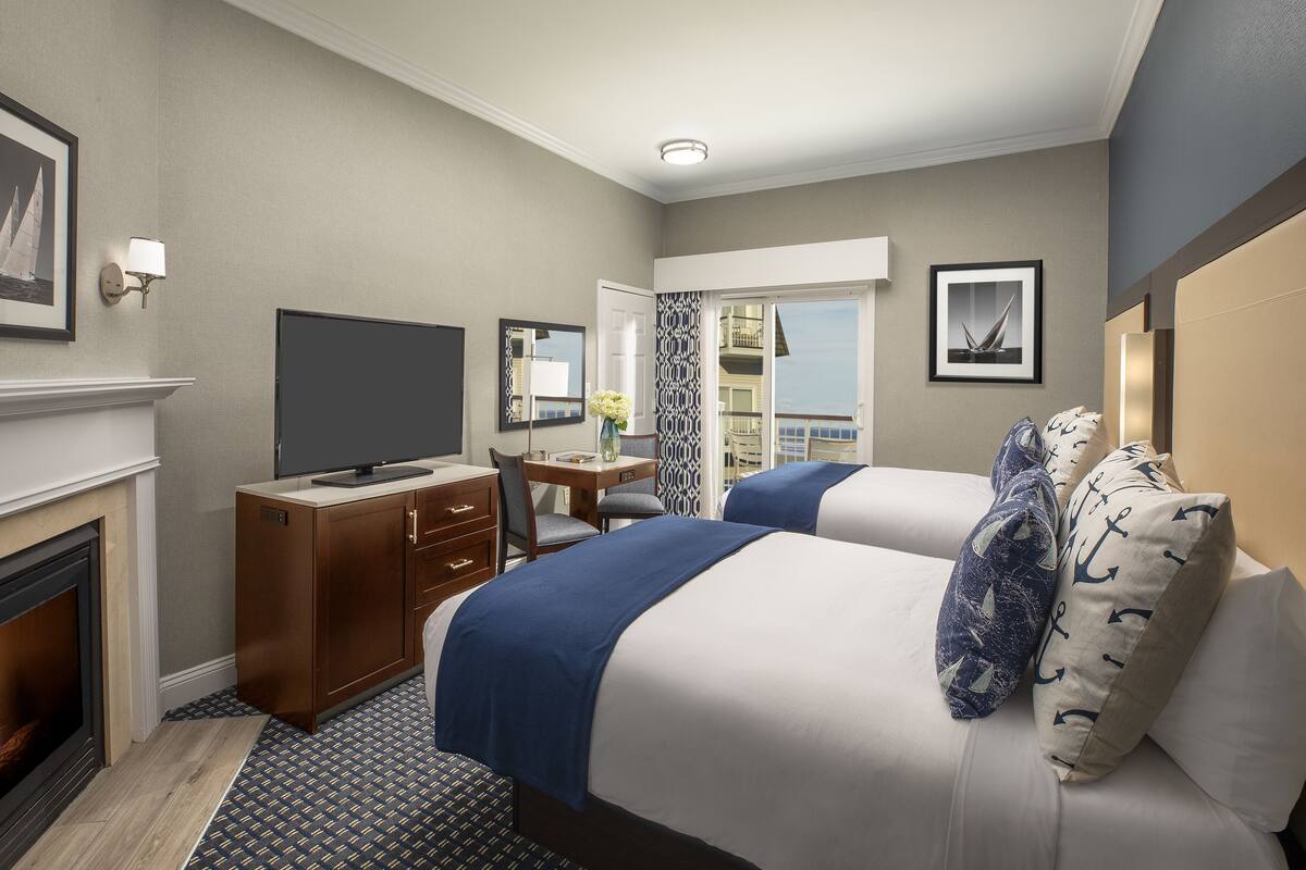 A hotel room with a fireplace, a television, a desk with a chair, two beds with matching pillows, and a balcony with a view.