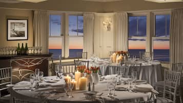 A dining area with tables set for a meal, candles, and a view of the ocean at sunset.
