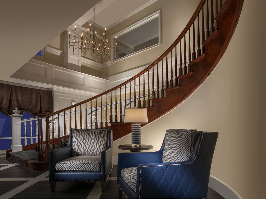 A grand staircase with a chandelier, two patterned armchairs, and a round table with a lamp.