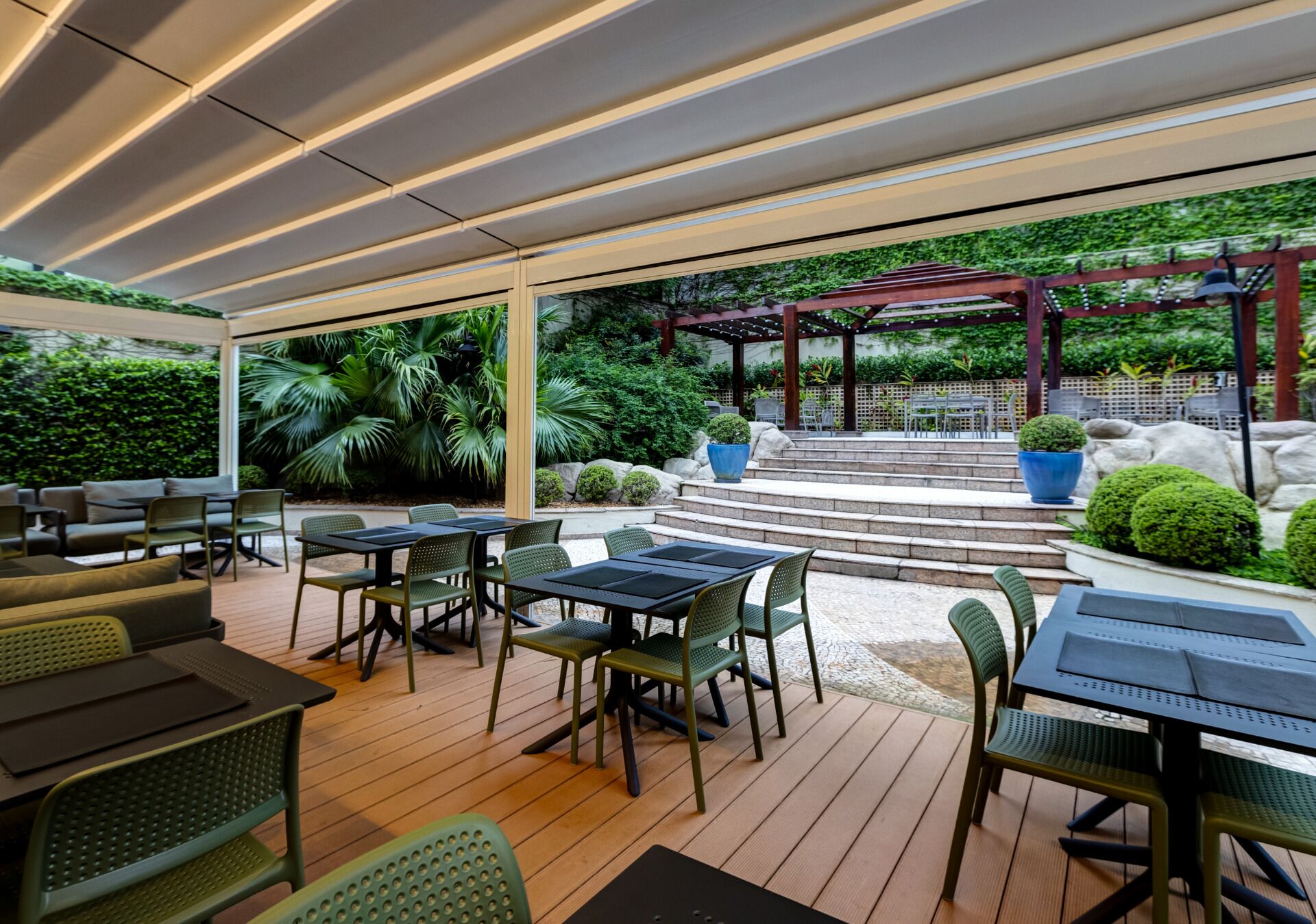 An outdoor patio with tables and chairs, a pergola, and a view of a garden.