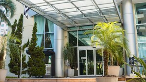 Una entrada de hotel moderna con puerta de cristal, palmeras y un paseo techado.