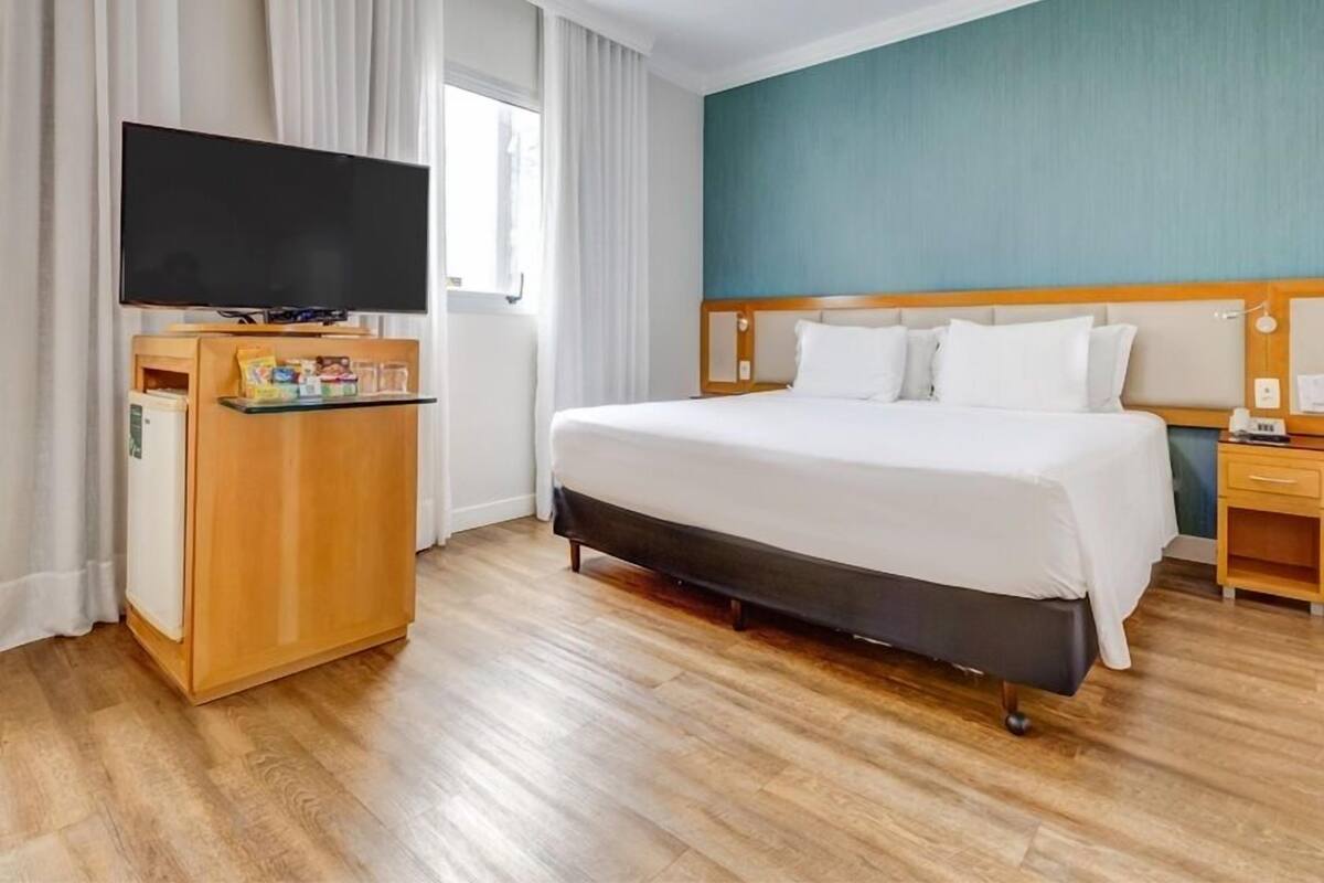 Ett hotellrum med en säng, en tv på ett träställ, ett skrivbord med en spegel och ett fönster med gardiner.