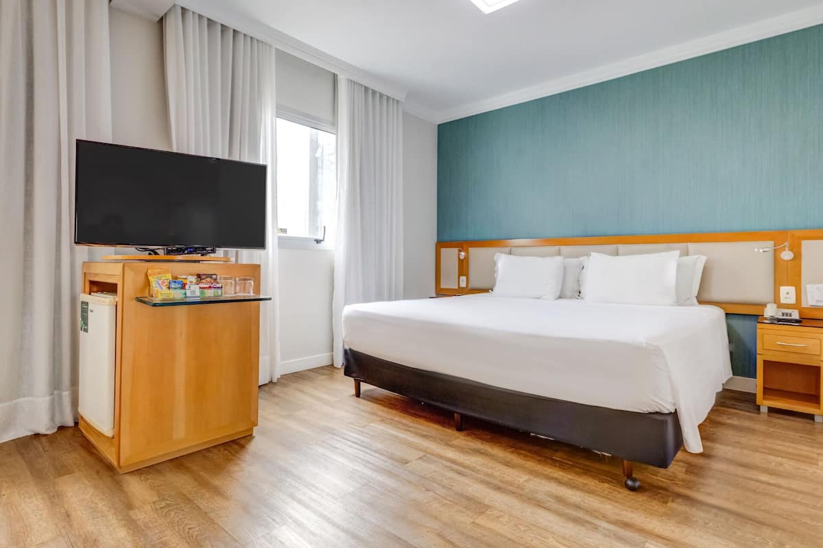 Ett hotellrum med en säng, en TV, en minibar och ett fönster med gardiner.