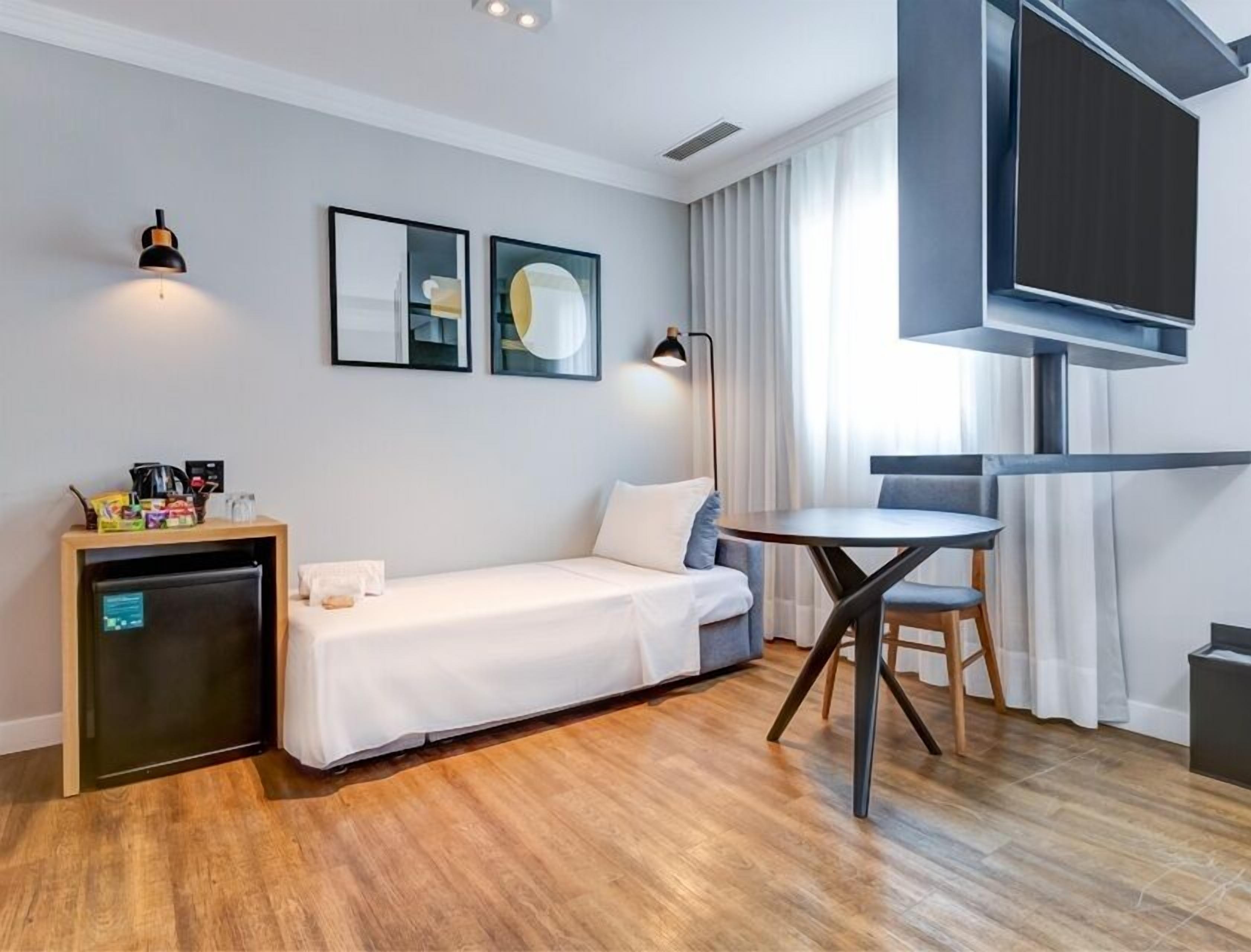 A modern hotel room with a bed, a small desk with a computer, a TV, and a window with curtains.