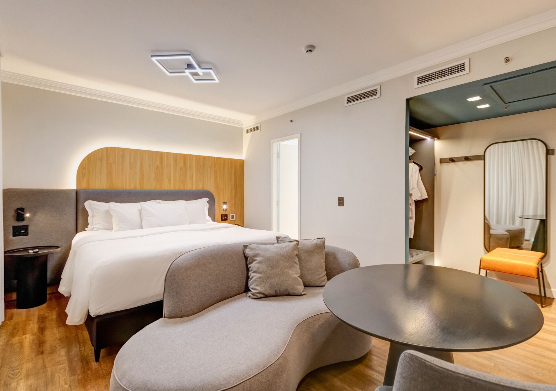 A modern hotel room with a curved sofa, a round table, a bed with white linens, and a wooden headboard.