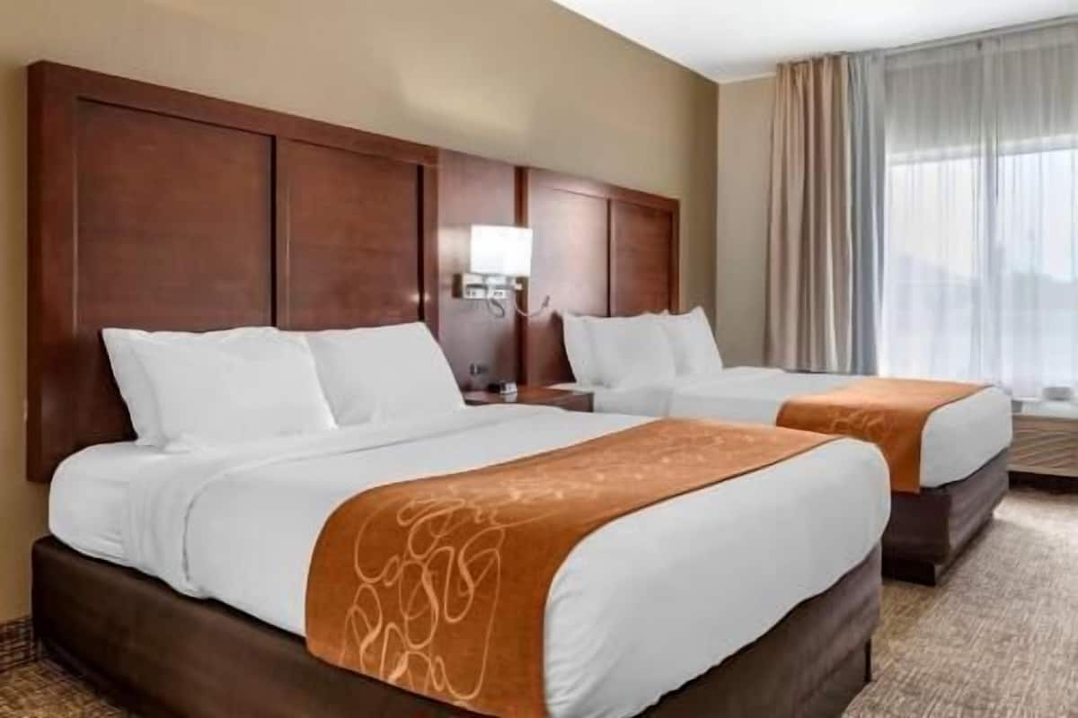 Habitación de hotel con dos camas, cabecero de madera, mesita de noche con lámpara y ventana con cortinas.
