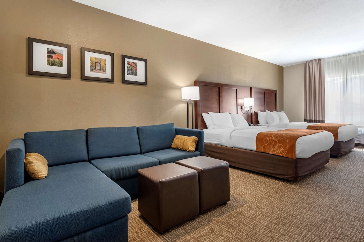 Habitación de hotel con un sofá azul, dos camas con cabeceros marrones, una mesita de noche de madera y tres cuadros enmarcados en la pared.