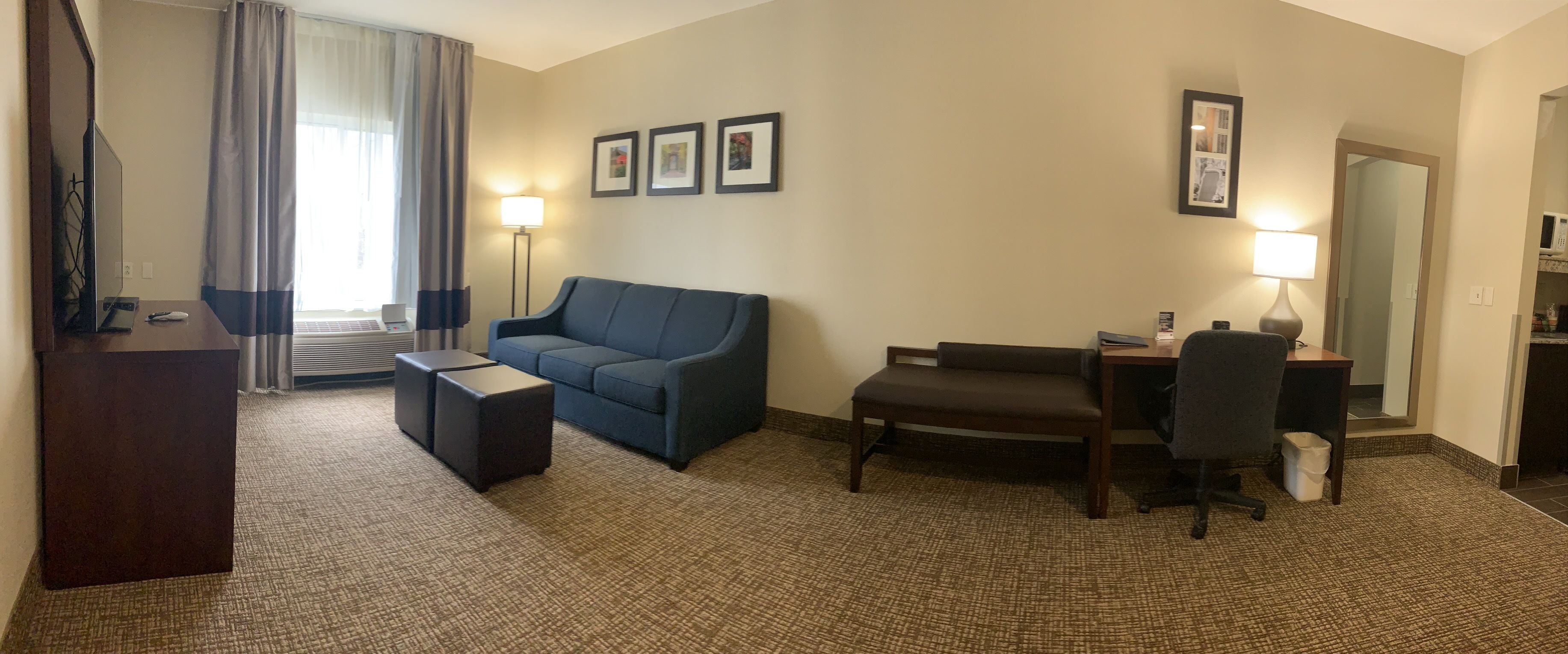 A hotel room with a blue sofa, a wooden desk, a chair, a small ottoman, and a TV. There are three framed pictures on the wall and a lamp on the desk.